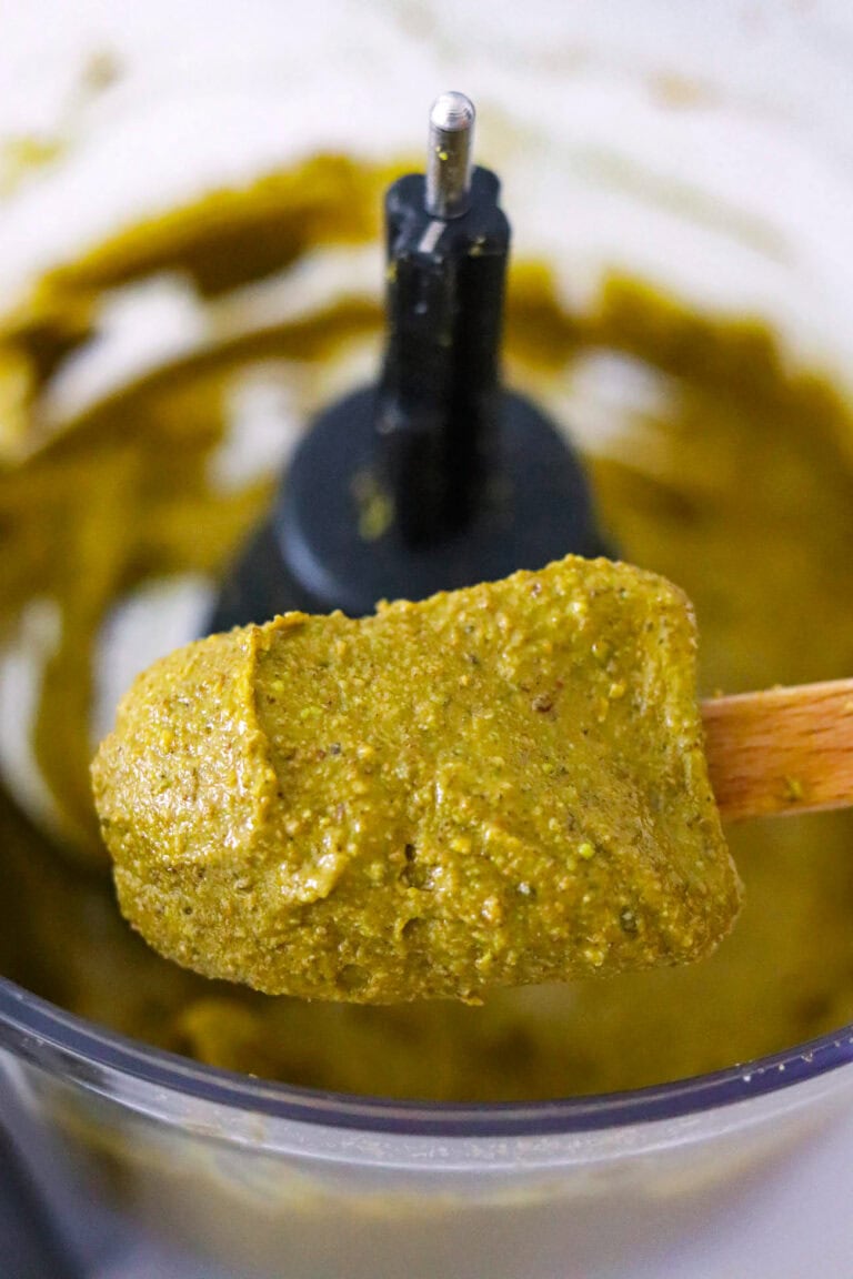 Mini silicone spatula with pistachio butter dripping down into a food processor bowl.