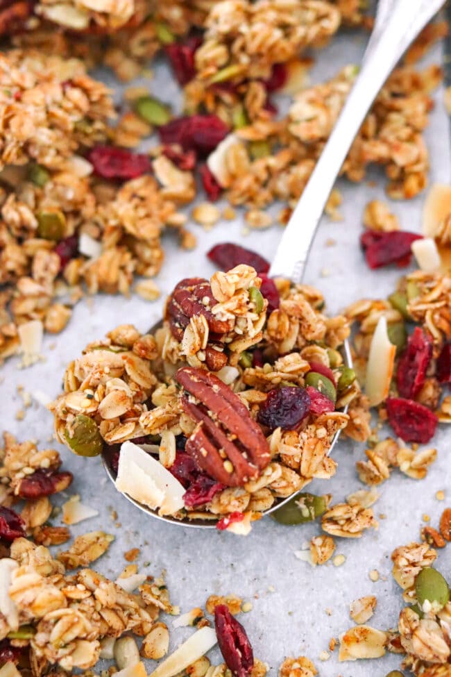 Closeup of spoon with pecan granola with dried cranberries and coconut flakes on a baking tray with granola.