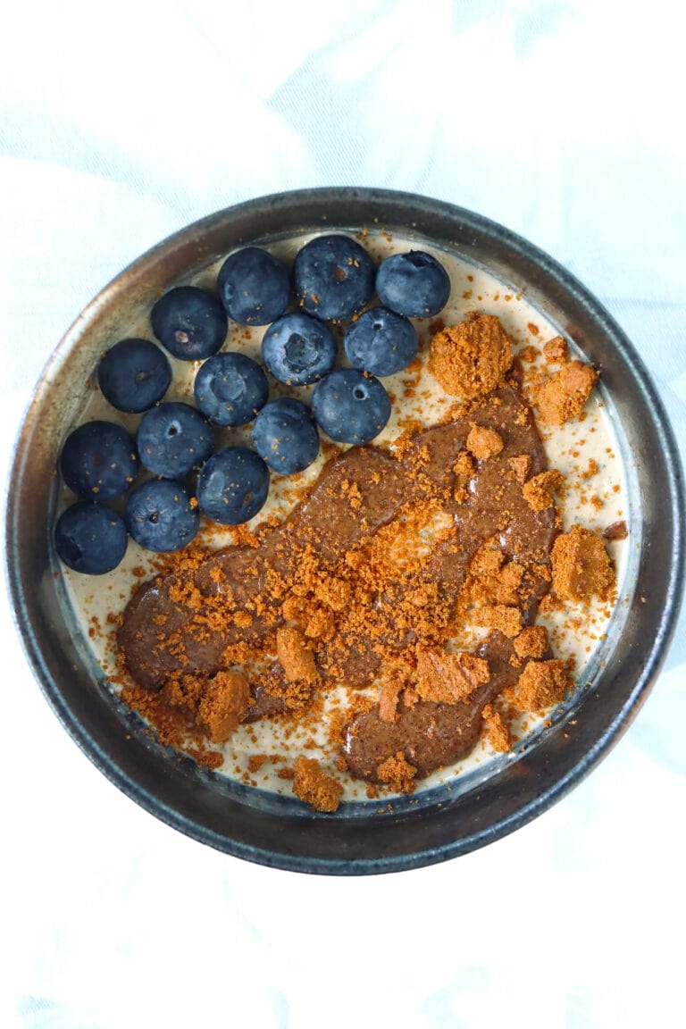 Gingerbread overnight oats in a black bowl topped with pecan butter, crumbled Biscoff cookie and blueberries.