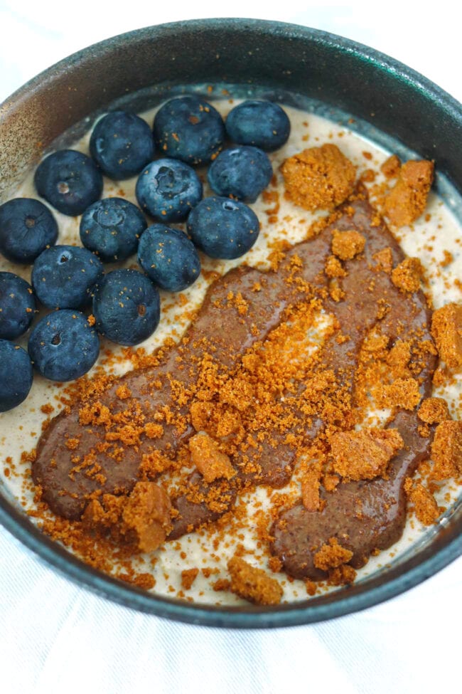 Closeup of bowl with gingerbread overnight oats topped with pecan butter, crumbled Biscoff cookie, and fresh blueberries.