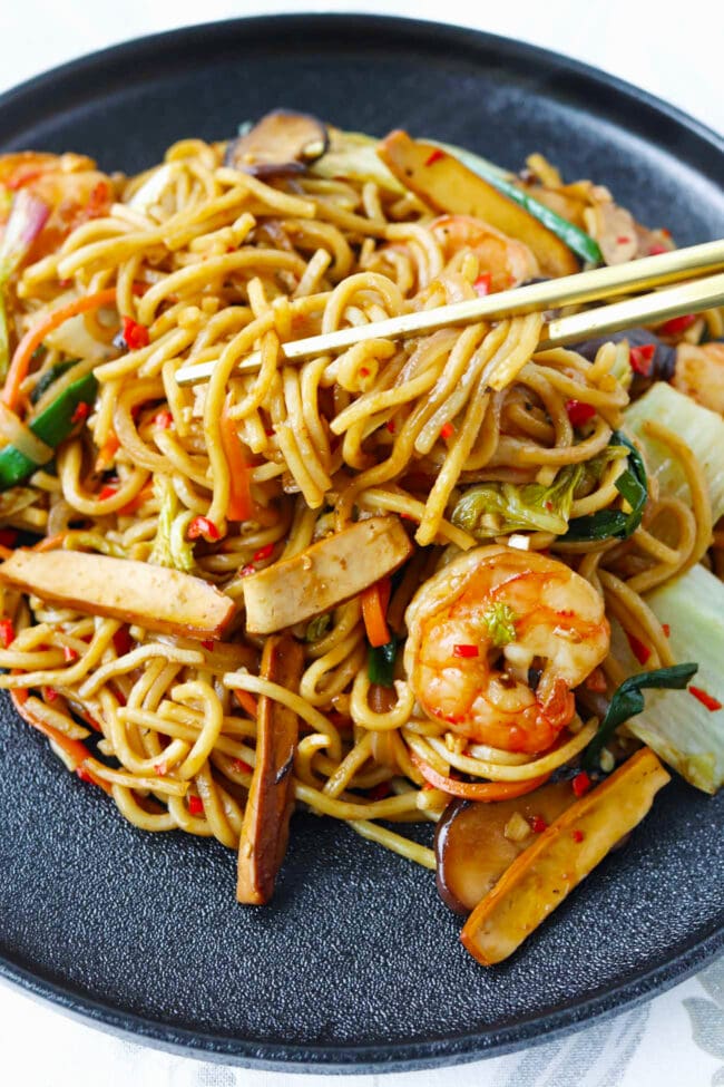 Chopsticks pulling up shrimp and tofu yakisoba noodles from plate.