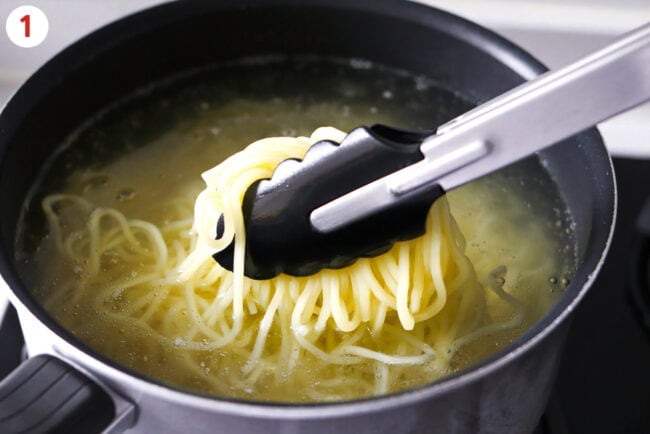 Tongs pulling up yakisoba noodles from pot of hot water.