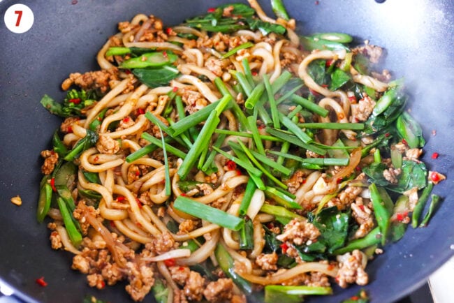 Added green onion to spicy pork noodles stir-fry in a wok.