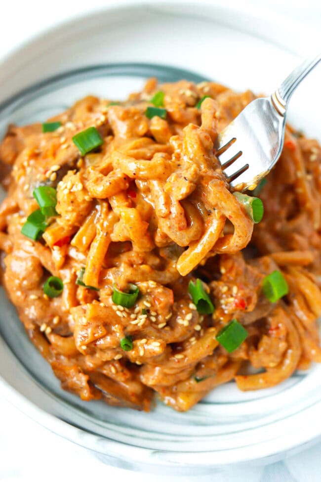 Fork pulling up a bite of creamy kimchi udon noodles with blended tofu sauce from a plate.