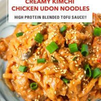Front view closeup of creamy kimchi noodles with chicken garnished with green onions and sesame seeds on a plate. Text overlay "Creamy Kimchi Chicken Udon Noodles", "High Protein Blended Tofu Sauce!!" and "That Spicy Chick".