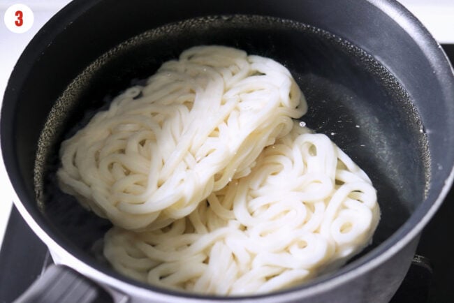 Frozen udon noodle cakes in a pot of boiled water.