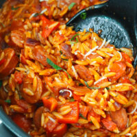 Closeup of spatula pushing spicy cajun tomato orzo with sausage and peppers to edge of skillet.