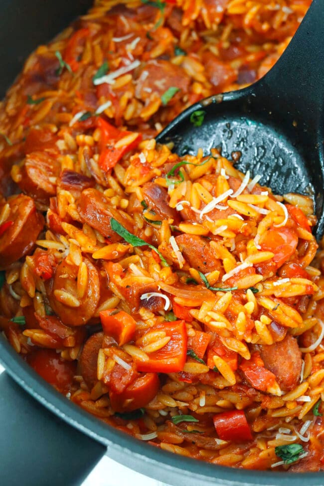Closeup of spatula pushing spicy cajun tomato orzo with sausage and peppers to edge of skillet.