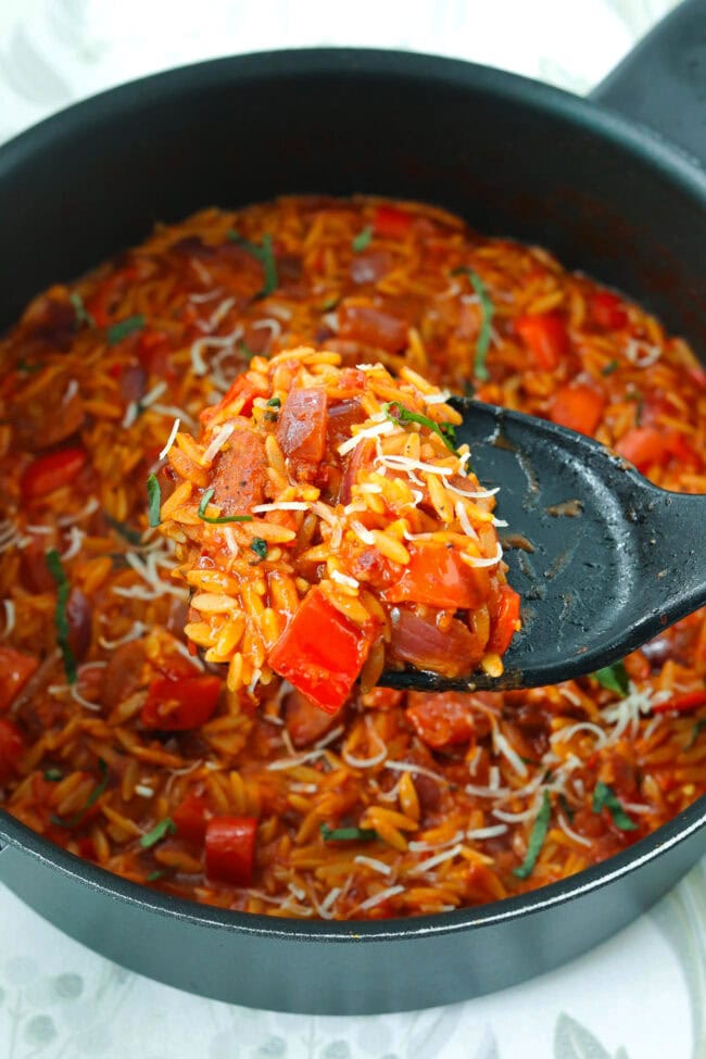 Spatula holding up a serving of cajun orzo with sausage, onion and peppers above pan.