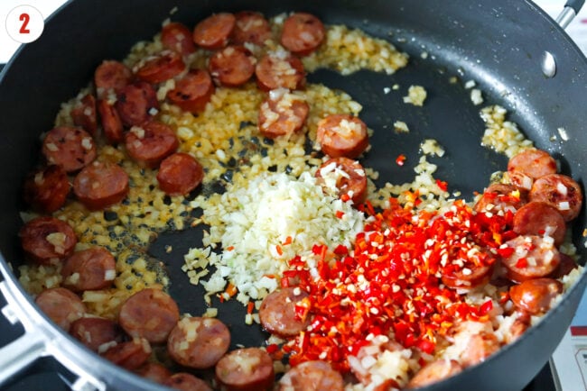 Minced garlic and red chilies in pan with sautรฉed sliced spicy sausage and yellow onion.