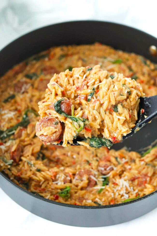 Spatula holding up a serving of spicy Italian sausage with orzo above pan.
