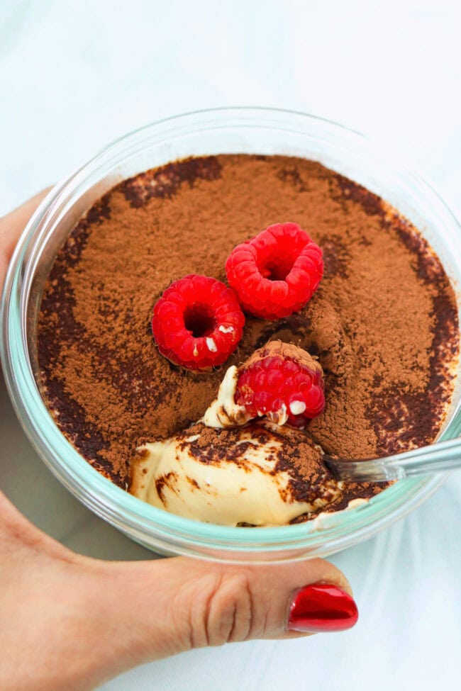 Hand holding small round dish with cottage cheese tiramisu and a spoon.