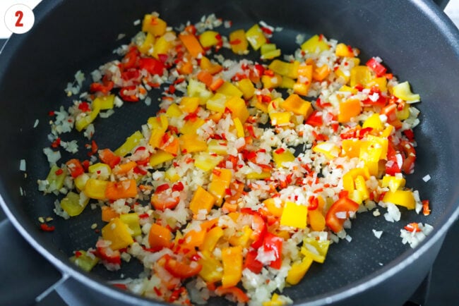 Sautéed yellow onion, red, yellow and orange bell peppers, garlic, and red chilies in a pan.