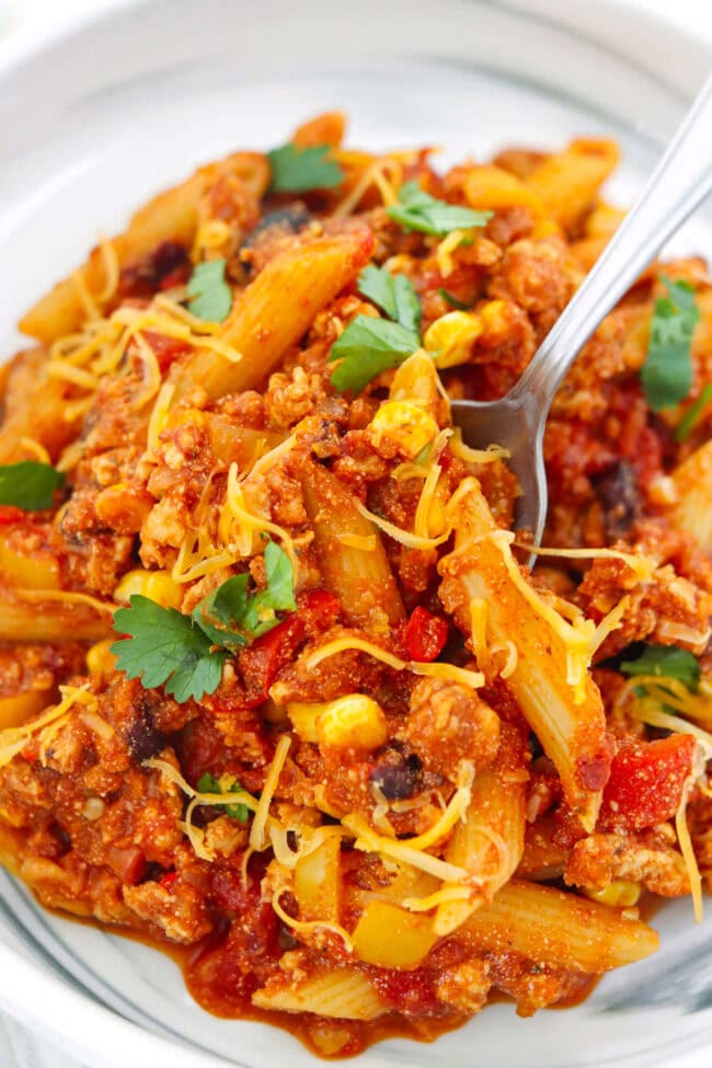Closeup of fork in a plate with chicken taco pasta topped with shredded cheddar and fresh cilantro.