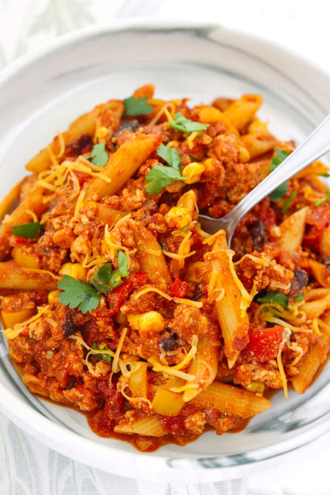 Fork in a plate with chicken taco pasta topped with shredded cheddar and fresh cilantro.