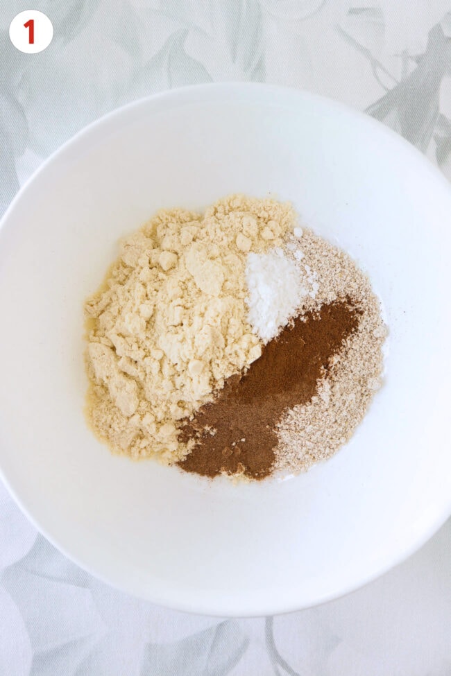 A white bowl with vanilla protein powder, blended oats, baking powder, ground cinnamon and apple pie spice.