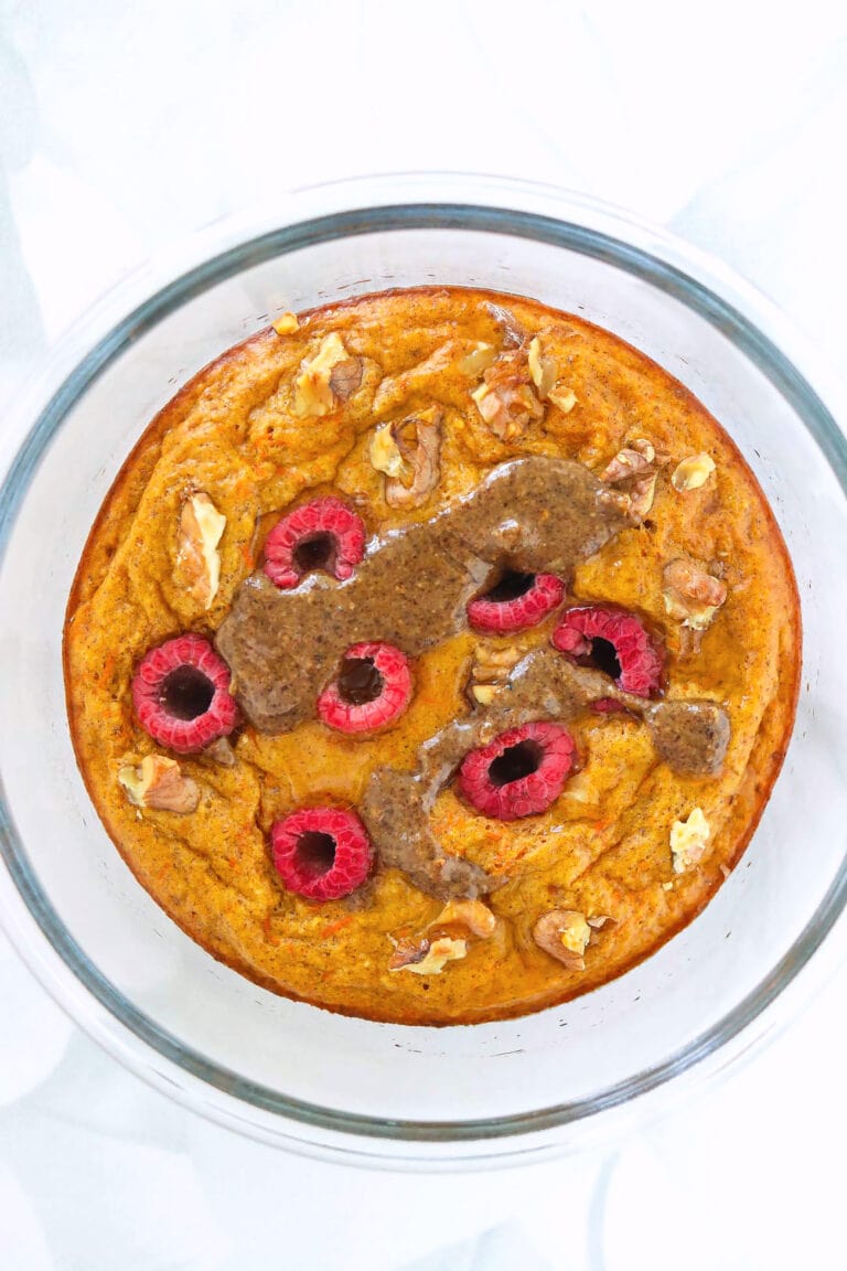 Carrot cake protein pancake in a round glass meal prep container topped with chopped walnuts, raspberries, pecan butter and maple syrup.