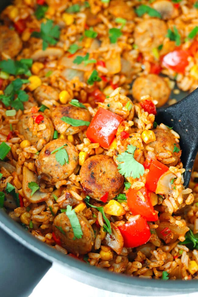 Closeup front view of spatula pushing rice and sausage with veggies to edge of pan.