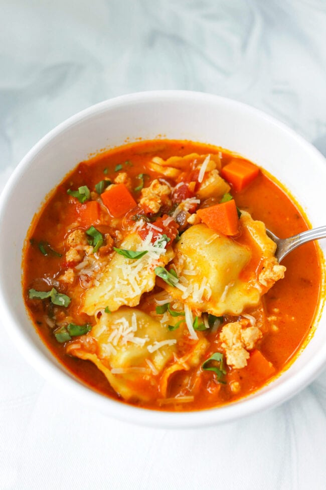 Spoon in bowl with spicy vodka ravioli soup with ground chicken topped with grated parmesan and sliced basil.