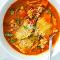 Spoon in bowl with spicy ravioli soup with ground chicken garnished with parmesan and basil.