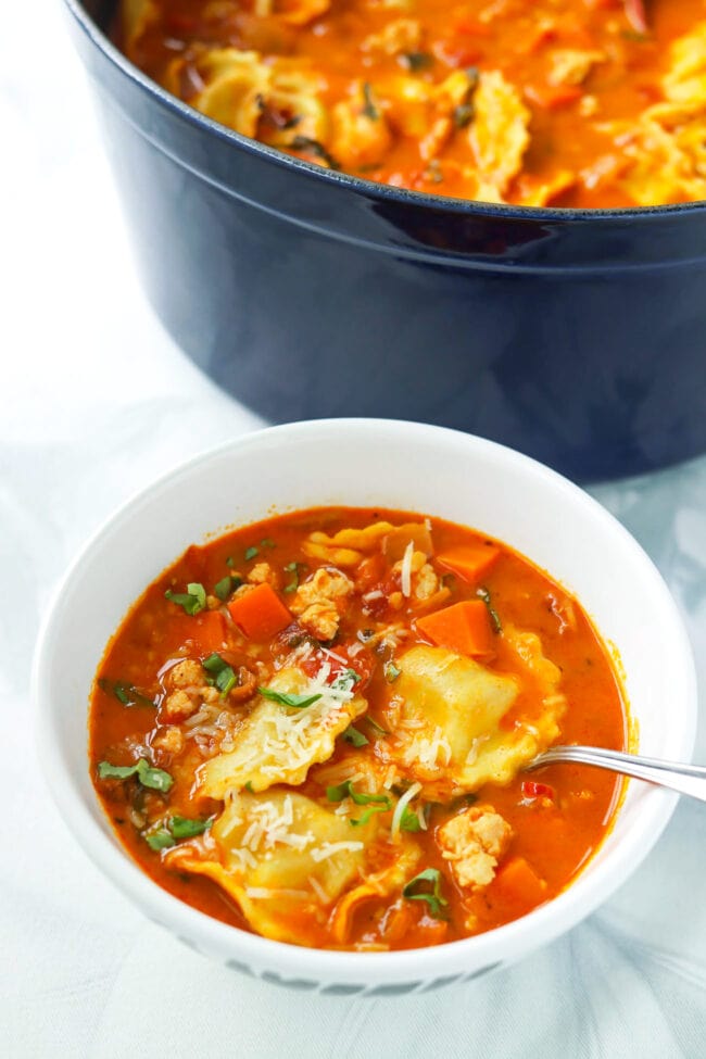 Front view of bowl and Dutch oven with spicy ravioli soup with ground chicken garnished with grated parmesan and sliced basil.