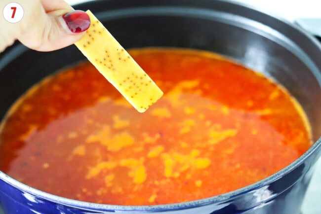 Hand holding a Parmigiano Reggiano cheese rind above a pot with soup.