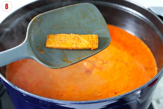 Softened parmigiano reggiano cheese rind on a spatula above a pot with soup.