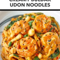 Closeup front view of spicy creamy Korean udon noodles with shrimp on a plate topped with black sesame seeds and green onions. Text overlay "Easy Weeknight Dinner!", "Creamy Buldak Udon Noodles" and "That Spicy Chick".
