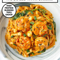 Plate with spicy creamy Korean udon noodles with shrimp topped with black sesame seeds and green onions. Text overlay "Creamy Buldak Udon Noodles with Shrimp", "High Protein Blended Tofu Sauce!" and "That Spicy Chick".