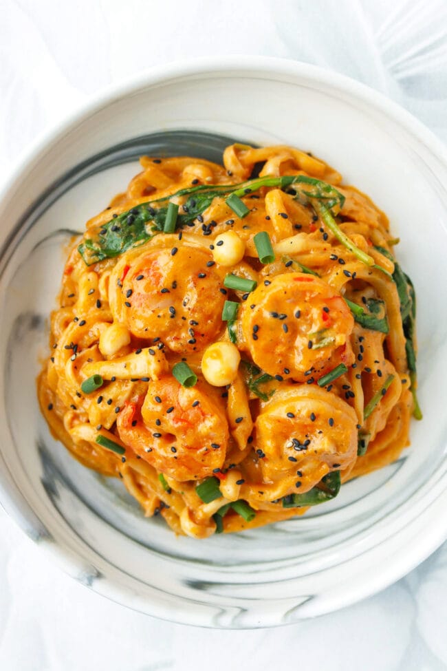 Closeup of plate with creamy tofu sauce buldak udon noodles with shrimp garnished with black sesame seeds and green onions.