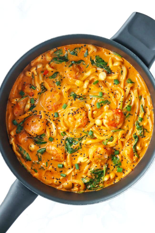Skillet with creamy tofu sauce buldak udon noodles with shrimp garnished with black sesame seeds and green onions.