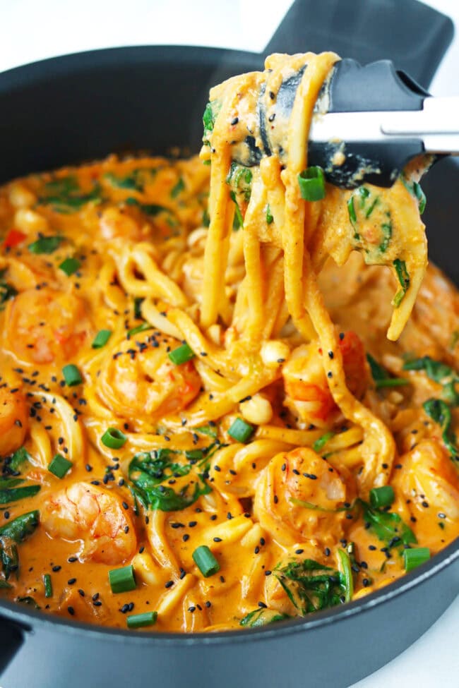 Tongs pulling up creamy buldak udon noodles with shrimp from a skillet.