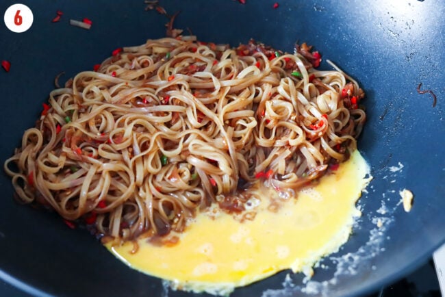 Noodles with side pushed to one side of wok and beaten egg cooking on other side.