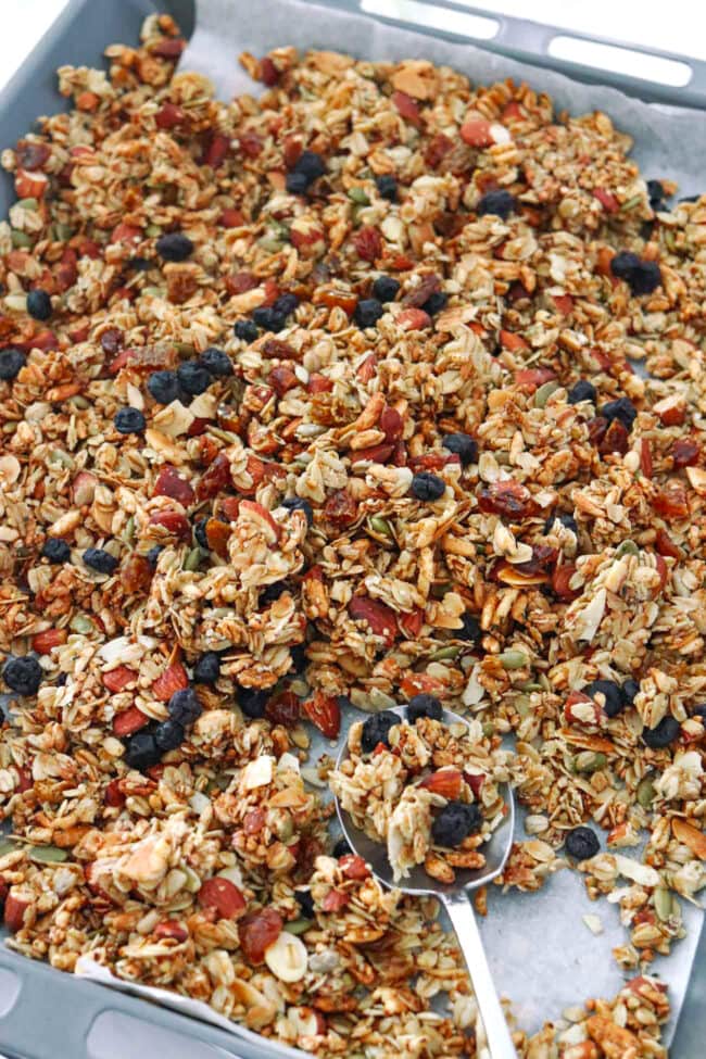 Vanilla almond granola on a baking tray with a spoon topped with dried blueberries and golden raisins.