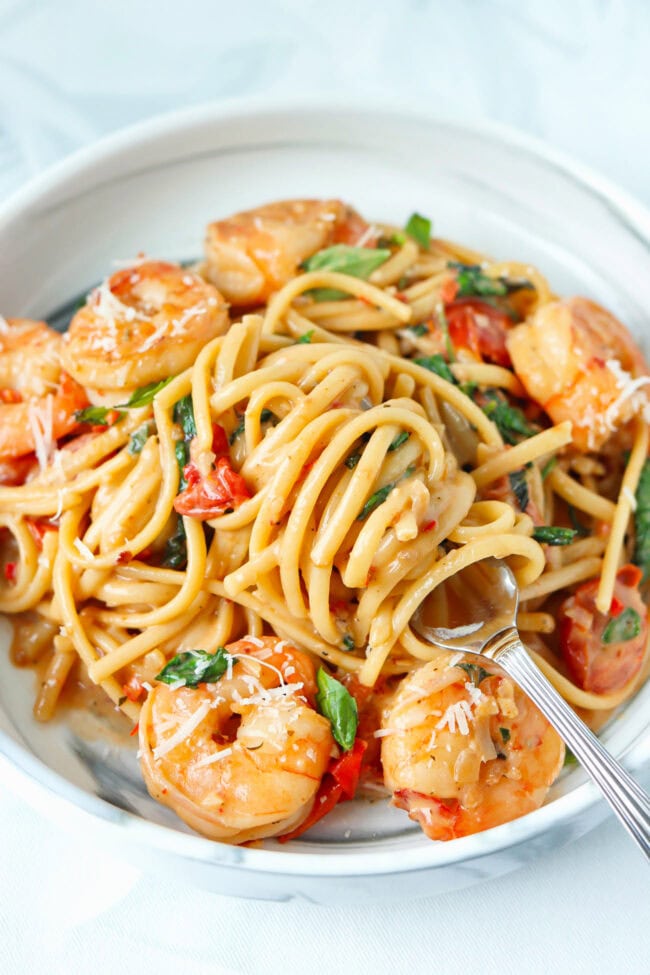Fork twirled into linguini noodles on a plate with creamy tomato shrimp pasta topped with parmesan and basil.