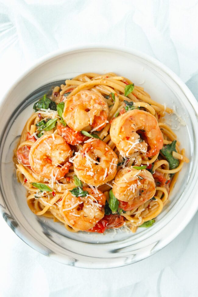 Closeup of white plate with creamy tomato shrimp pasta with cherry tomatoes, spinach, and basil topped with grated parmesan.