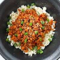 Spicy Korean ground chicken topped with sesame seeds and green onions on steamed rice in a black bowl.