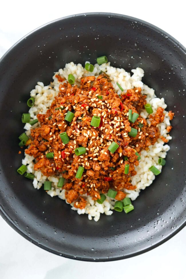 Spicy Korean ground chicken topped with sesame seeds and green onions on steamed rice in a black bowl.