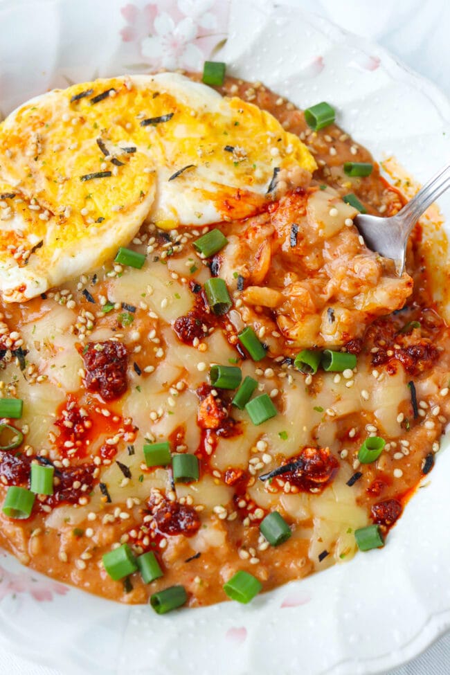 Spoon in a plate of savory oats with kimchi topped with cheese, fried egg, furikake, green onions, and chili crisp.