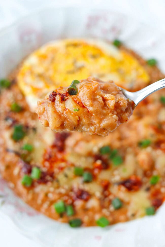 Spoon holding up a bite of savory oatmeal with kimchi above a plate.
