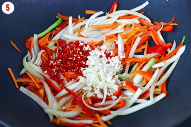 Garlic and red chilies in a wok with stir-fried sliced onion, carrots and spring onion bulb parts.