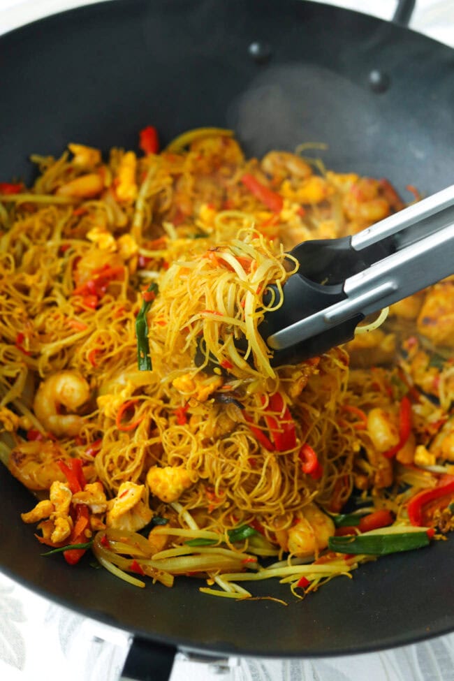 Tongs holding up stir-fried curry noodles with chicken, shrimp, egg and vegetables from a wok with noodles.