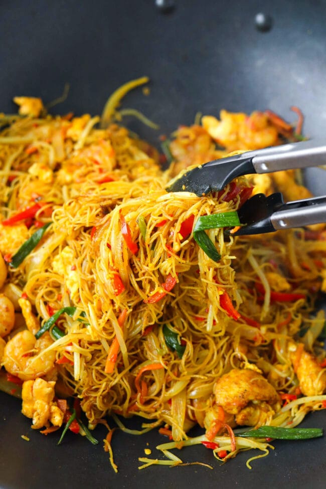 Tongs holding up stir-fried Singapore Noodles with chicken, shrimp, egg and vegetables from a wok with noodles.