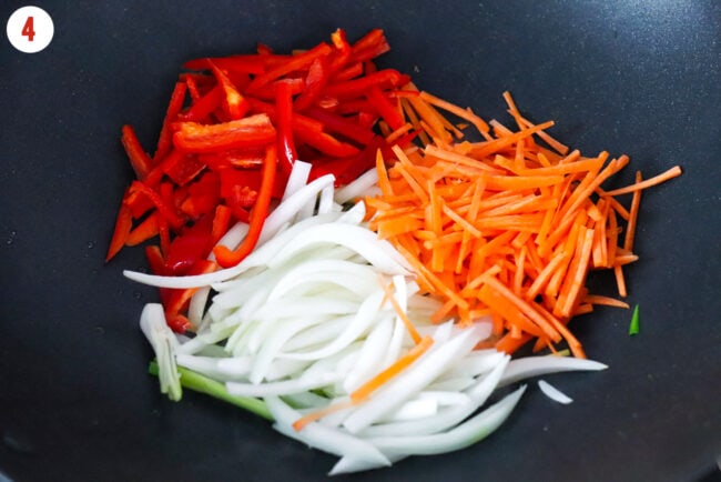 Sliced carrots, onion, spring onion white and light green parts, and red bell pepper in wok.