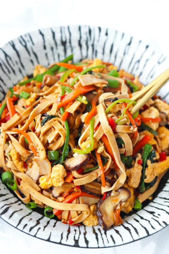 Chopsticks pulling out tofu noodles stir-fry with vegetables from a plate.