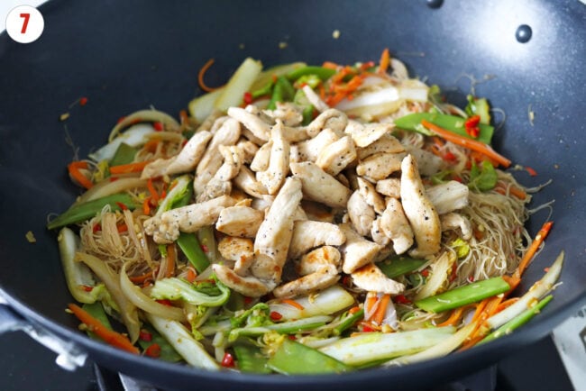 Cooked chicken pieces in a wok with stir-fried vegetables and thin rice vermicelli noodles.