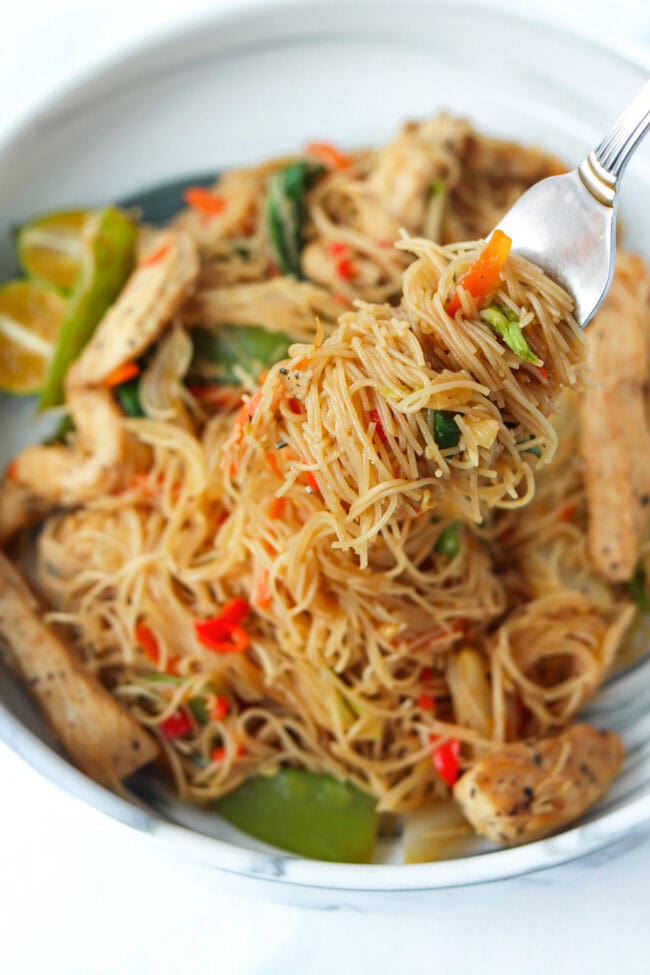 Fork with rice vermicelli noodles and chicken stir-fry held above a plate with pancit bihon.