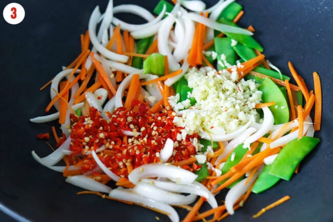 Minced garlic and fresh red chilies in a wok with stir-fried onion, snow peas, and thinly sliced carrots.
