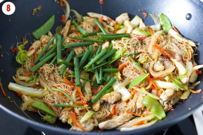 Spring onion, black pepper in a wok with stir-fried chicken and thin rice vermicelli noodles.