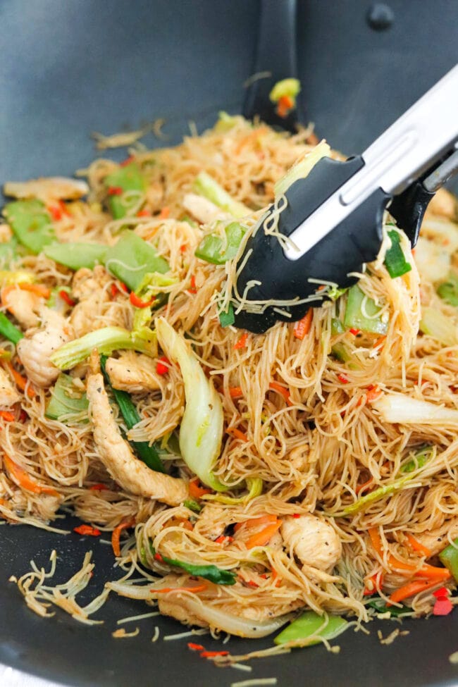 Tongs holding up a bunch of stir-fried chicken with vermicelli noodles, vegetables, and green onions in a wok.